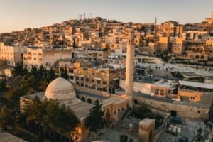 Aerial view of a historic Middle Eastern city at sunset, featuring a domed mosque and a tall minaret among sandstone buildings.
