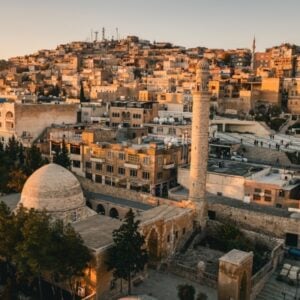Aerial view of a historic Middle Eastern city at sunset, featuring a domed mosque and a tall minaret among sandstone buildings.