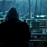 Hooded figure at a workstation with multiple monitors displaying code in a dark data center, suggesting a hacking scene.