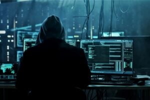 Hooded figure at a workstation with multiple monitors displaying code in a dark data center, suggesting a hacking scene.