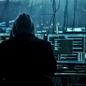 Hooded figure at a workstation with multiple monitors displaying code in a dark data center, suggesting a hacking scene.