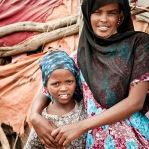 Woman in a colorful dress hugs a young girl wearing a blue patterned headscarf, outdoors with wooden poles and fabric behind them.
