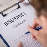Close-up of a hand writing on an insurance claim form clipped to a blue clipboard.