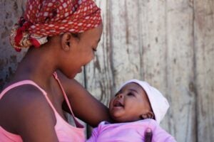 Older sister with a red headscarf cradles a smiling baby in pink/purple, against a wooden wall.