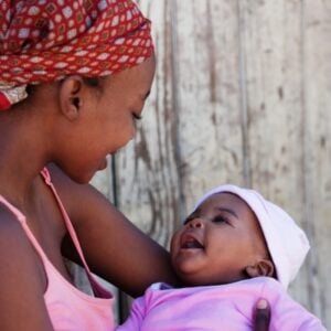 Older sister with a red headscarf cradles a smiling baby in pink/purple, against a wooden wall.
