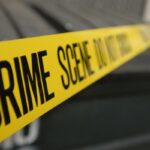 Yellow crime scene tape reading 'CRIME SCENE DO NOT ENTER' stretched across a scene, with a blurred background vehicle behind it.