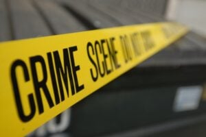 Yellow crime scene tape reading 'CRIME SCENE DO NOT ENTER' stretched across a scene, with a blurred background vehicle behind it.