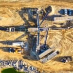 Aerial view of a construction site with a long conveyor system, concrete structures, and parked trucks on dusty yellow ground.
