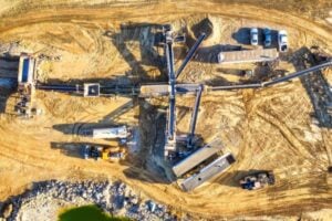Aerial view of a construction site with a long conveyor system, concrete structures, and parked trucks on dusty yellow ground.