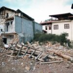 Rubble-strewn street with a partially destroyed two-story building and exposed wooden beams amid debris in a ruined neighborhood.