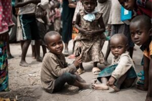 Group of children outdoors; two smiling kids sit on the dirt foreground while others stand around them on a dusty ground.