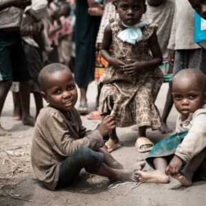 Group of children outdoors; two smiling kids sit on the dirt foreground while others stand around them on a dusty ground.