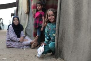 Girl in teal, crouching by a concrete wall and holding a colorful booklet; others sit nearby in a dusty doorway of a corrugated settlement.