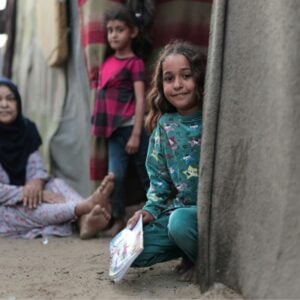 Girl in teal, crouching by a concrete wall and holding a colorful booklet; others sit nearby in a dusty doorway of a corrugated settlement.