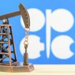 Miniature oil pump jack model on a wooden surface with a blue backdrop and white shapes in the background.