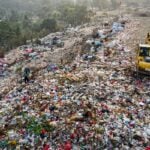 A vast landfill with piles of trash and a yellow excavator sorting waste on a hilltop, with scattered bags and debris across the site.