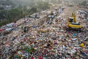 A vast landfill with piles of trash and a yellow excavator sorting waste on a hilltop, with scattered bags and debris across the site.