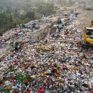 A vast landfill with piles of trash and a yellow excavator sorting waste on a hilltop, with scattered bags and debris across the site.