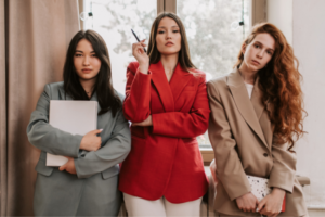 Three young women stand in a hallway, posing confidently; center woman in red blazer holds a pen, left holds a folder, right in beige blazer.