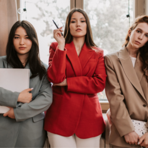 Three young women stand in a hallway, posing confidently; center woman in red blazer holds a pen, left holds a folder, right in beige blazer.
