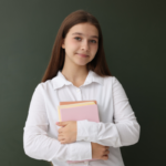 Student with long brown hair in a white shirt holds pink and orange notebooks in front of a chalkboard, smiling softly.