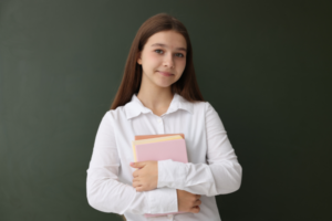 Student with long brown hair in a white shirt holds pink and orange notebooks in front of a chalkboard, smiling softly.