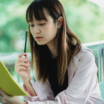 Young woman sits outdoors, reading a yellow notebook and holding a pencil, absorbed in study.