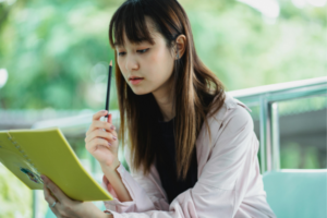 Young woman sits outdoors, reading a yellow notebook and holding a pencil, absorbed in study.
