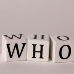 Three white letter blocks arranged on a pale surface spell 'WHO' (W, H, O).