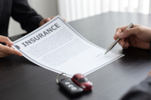 Two people sign an insurance contract at a desk, with car keys placed nearby on the table.