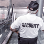 Security guard on an escalator, viewed from behind, wearing a white shirt with 'SECURITY' on the back.