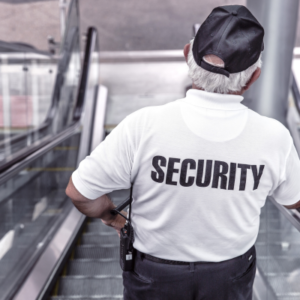 Security guard on an escalator, viewed from behind, wearing a white shirt with 'SECURITY' on the back.