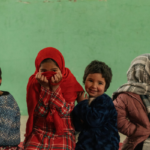 Group of smiling children sitting in a row against a green wall; a girl in a red hijab covers her mouth with her hands.