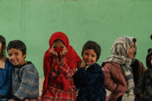 Group of smiling children sitting in a row against a green wall; a girl in a red hijab covers her mouth with her hands.