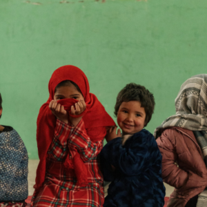 Group of smiling children sitting in a row against a green wall; a girl in a red hijab covers her mouth with her hands.