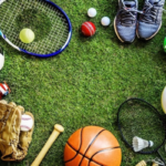 Assortment of sports gear on a grass field: soccer ball, basketball, volleyball, tennis racket, baseball bat and glove, shuttlecock, rugby ball, sneakers, and other equipment.