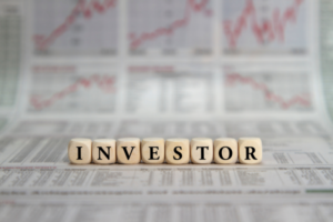 Wooden letter blocks spell INVESTOR on a newspaper with stock charts in the background.