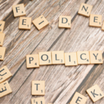 Scrabble tiles spell 'POLICY' on a wooden table, with scattered letters around.