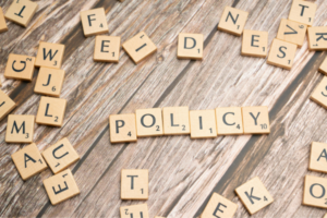 Scrabble tiles spell 'POLICY' on a wooden table, with scattered letters around.