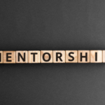 Wooden letter blocks spell 'MENTORSHIP' on a dark surface, illustrating the concept of mentorship.
