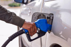 Person refueling a silver car with a blue gas pump nozzle at the fuel inlet in a driveway or station setting.