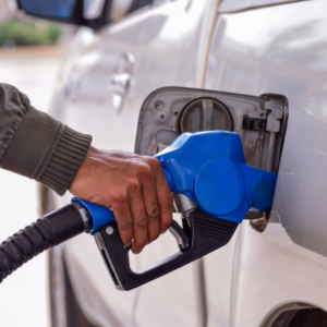 Person refueling a silver car with a blue gas pump nozzle at the fuel inlet in a driveway or station setting.