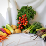 Assorted fresh vegetables arranged on white fabric: tomatoes, greens, corn, carrots, peppers, potatoes, cucumbers, beets, zucchini, and eggplant.