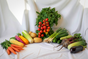 Assorted fresh vegetables arranged on white fabric: tomatoes, greens, corn, carrots, peppers, potatoes, cucumbers, beets, zucchini, and eggplant.