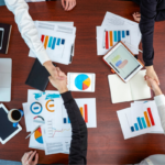 Overhead view of colleagues shaking hands across a conference table cluttered with charts, tablets, and laptops, signaling agreement after a data review.
