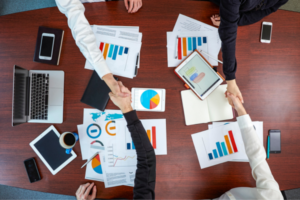 Overhead view of colleagues shaking hands across a conference table cluttered with charts, tablets, and laptops, signaling agreement after a data review.