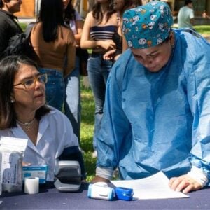 Female healthcare worker in a white coat sits at a table with medical supplies; another clinician in blue scrubs and a scrub cap leans over to review papers outdoors at a community health event, with bystanders in the background.