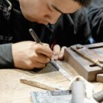 Close-up of a craftsman’s hands engraving a metal strip on a wooden workbench with engraving tool and nearby tools nearby