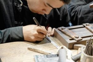Close-up of a craftsman’s hands engraving a metal strip on a wooden workbench with engraving tool and nearby tools nearby