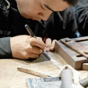 Close-up of a craftsman’s hands engraving a metal strip on a wooden workbench with engraving tool and nearby tools nearby